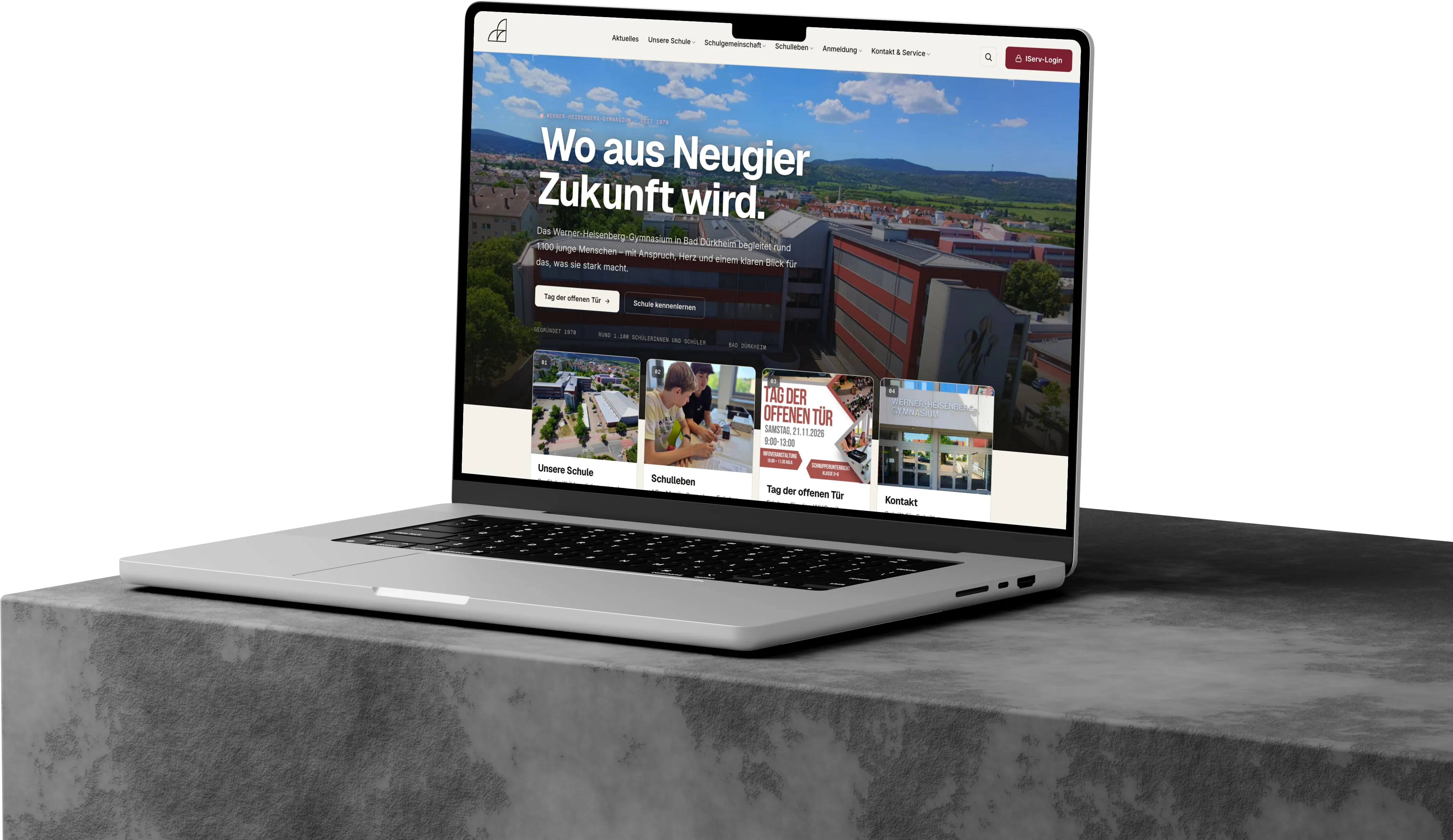 MacBook auf einem Betonsockel mit der Startseite des Werner-Heisenberg-Gymnasiums – „Wo aus Neugier Zukunft wird.“